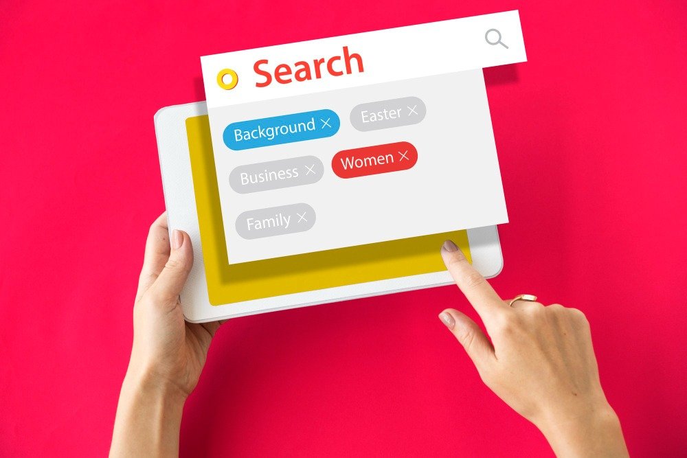 Hands holding a digital tablet against a pink background showing a search bar with tag filters, representing metadata optimization and technical multilingual SEO.