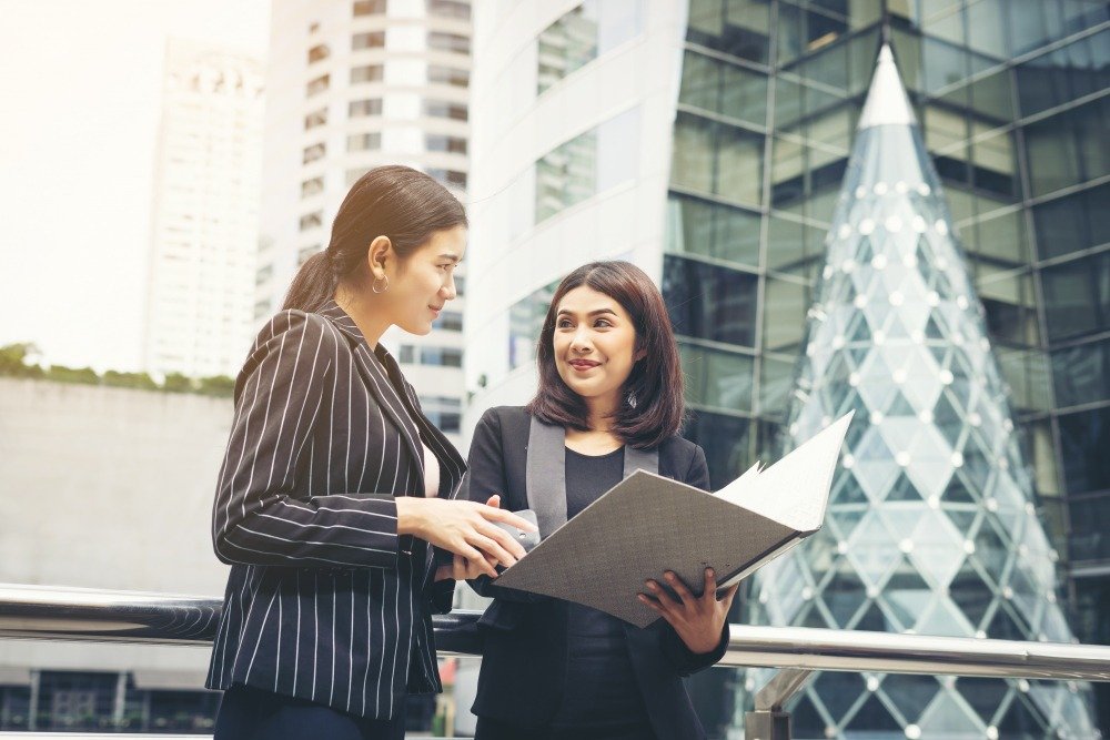 Two business professionals discussing documents outside a modern skyscraper, representing B2B partnerships and certified translation services in Malaysia